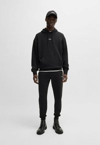 Czarna bluza z kapturem z kieszenią z przodu i logo, w zestawie z pasującymi spodniami dresowymi i czarnymi butami na sznurówki. Dodatkiem jest czarna czapka.