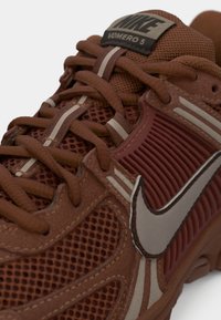 Barna Nike Vomero 5 futócipő, hálós felsőrésszel, párnázott fűzőkkel és fényvisszaverő swoosh logóval az oldalon.