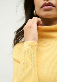 Frau, die den Kragen eines gelben gerippten Rollkragensweaters mit Kabelstrick-Ärmeldetails berührt und einen goldenen Creolenohrring trägt.