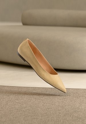 Beige spiss tå flat sko med svart såle svevende over et teksturert beige teppe, med en beige polstret sofa i bakgrunnen.