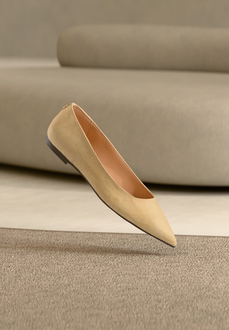 Chaussure plate beige à bout pointu avec une semelle noire flottant au-dessus d'un tapis beige texturé, avec un canapé beige rembourré en arrière-plan.