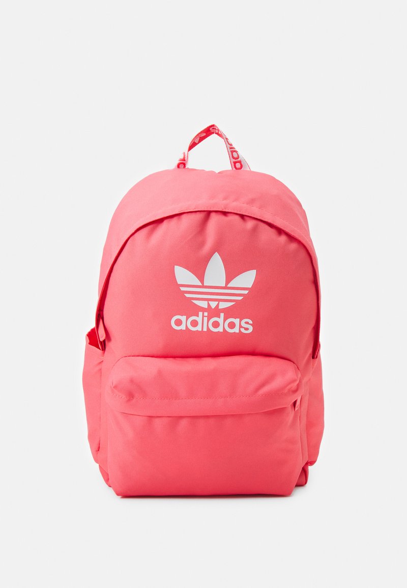 Rožinė Adidas kuprinė su apvalia forma, pagaminta iš audinio, su priekinėmis kišenėmis ir baltu logotipu, ant kurio pavaizduotos trys juostelės.