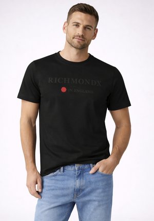 Uomo che indossa una T-shirt nera RichmondX con grafica di una rosa rossa e jeans blu chiaro, in piedi davanti a uno sfondo bianco uniforme.