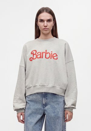 Kvinde med langt brunt hår iført en lysegrå sweatshirt med rød "Barbie" tekst og løstsiddende blå jeans, stående mod en ensfarvet hvid baggrund.