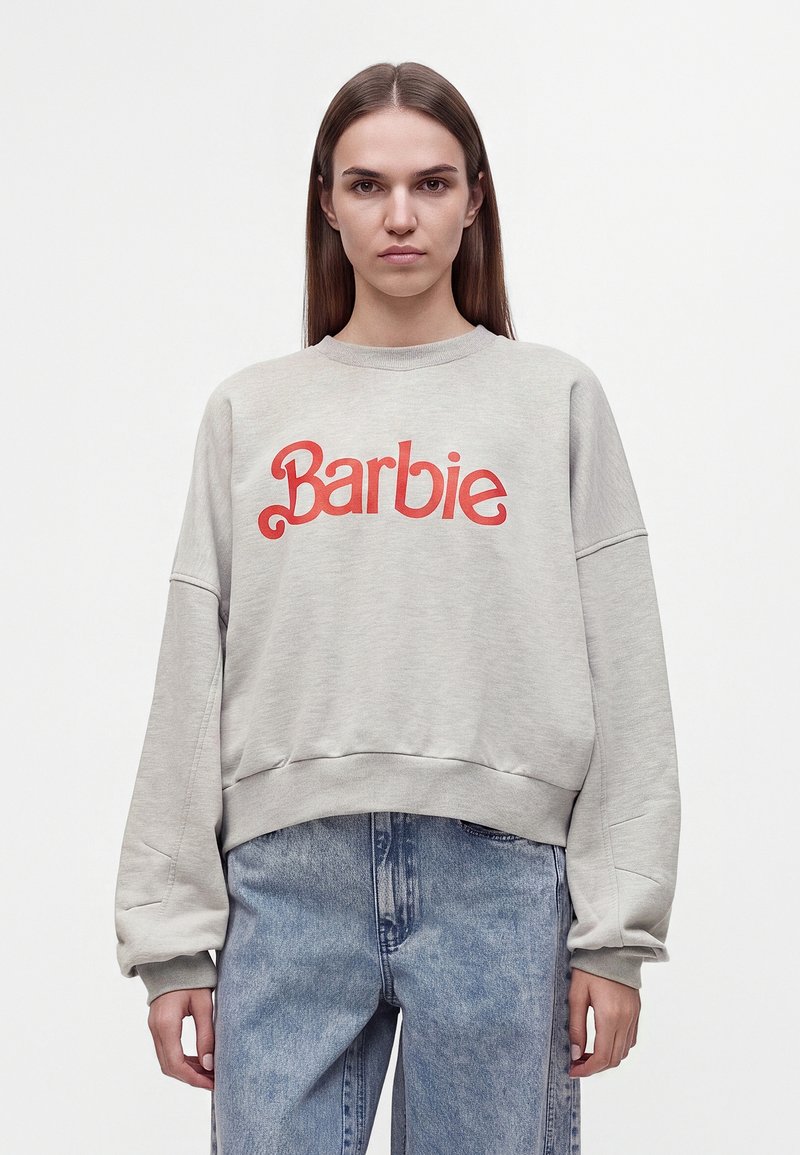 Femme aux longs cheveux bruns portant un sweat-shirt gris clair avec un texte rouge "Barbie" et un jean bleu large, debout sur un fond blanc uni.