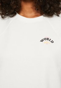 Sudadera blanca con cuello redondo y mangas raglán. Presenta un gráfico multicolor "SURFING WORLD ROXY" en el pecho. Textura suave.