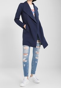 Personne portant un manteau bleu marine à ouverture frontale, un t-shirt blanc avec un graphique, un jean skinny bleu clair déchiré et des baskets blanches, se tenant avec une main dans la poche.