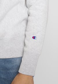 Grauer Sweatshirt mit geripptem Bündchen, Fleece-Textur und kleinem gestickten Logo auf dem Ärmel. Kombiniert mit Jeans.