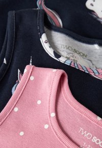 Dve detské body bez rukávov, jedno ružové s bielymi bodkami a jedno tmavomodré s motívom jednorožca, uložené na sivom povrchu.