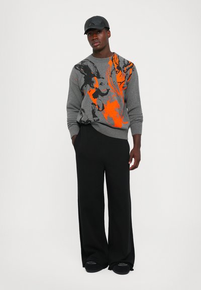 Grau gestrickter Pullover mit abstrakten Mustern in Schwarz und Orange, kombiniert mit schwarzen, weiten Hosen. Die Mütze hat eine strukturierte Oberfläche.