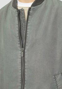 Szara kurtka bomberka o gładkim, matowym wykończeniu, z zapięciem na zamek, prążkowanym dekoltem i bocznymi kieszeniami. Charakteryzuje się lekką teksturą materiału.