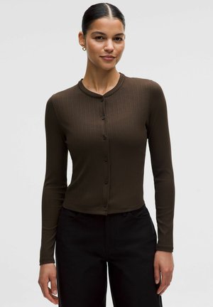 Ung kvinde med mørkt, tilbageglattet hår iført en figursyet brun ribstrikket cardigan med knapper og sorte bukser, stående mod en ensfarvet baggrund.