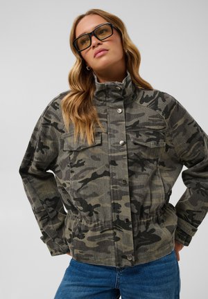 Femme portant de grandes lunettes de soleil et une veste camouflage verte avec des boutons, un jean bleu, debout les mains sur les hanches.