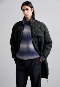 Veston bomber matelassé vert foncé avec deux poches avant et une fermeture éclair. Pull dégradé doux bleu et gris en dessous, associé à un pantalon sombre.