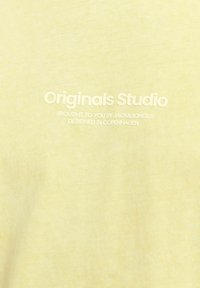 Šviesiai geltona medvilninė marškinė su švelniu paviršiumi. Puošia balta spaudinė su tekstu: "Originals Studio" ir prekės ženklo informacija subtiliu, švariu šriftu.
