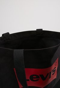 Černá plátěná taška s červeným logem Levi's, která má dvojité černé látkové uchy a neupravené horní otvory.