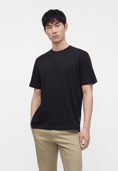 Homme aux cheveux courts noirs portant un t-shirt noir uni à manches courtes et un pantalon beige, debout devant un fond blanc.
