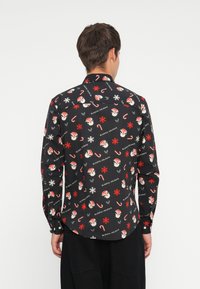 Chemise noire à manches longues ornée d'un motif de fête avec des visages de Santa, des cannes en sucre d'orge, des flocons de neige et le texte "Joyeuses Fêtes".