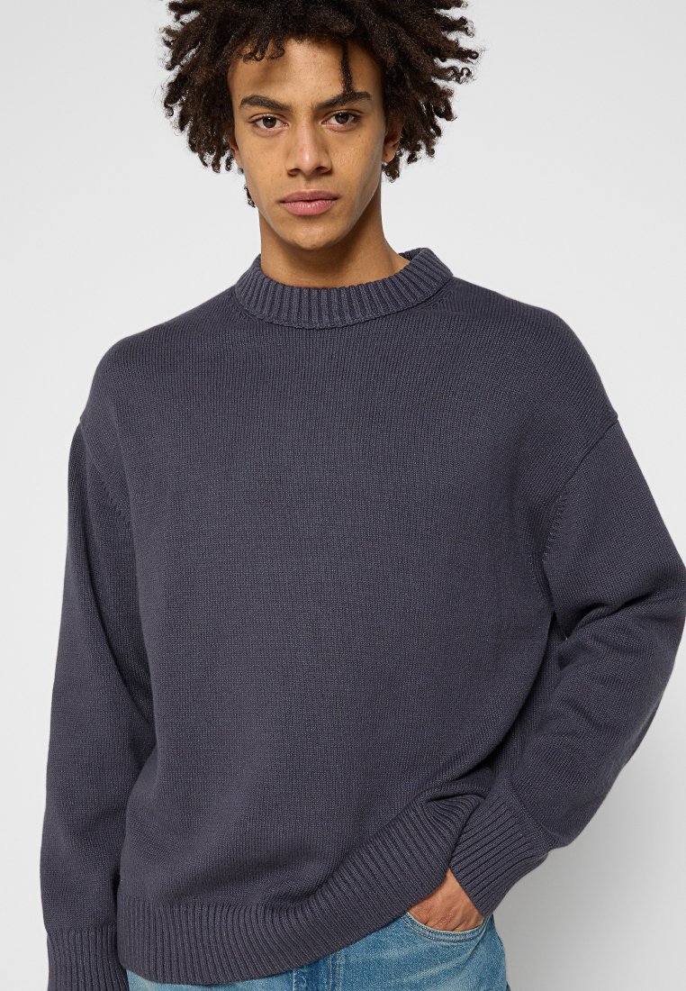 Giovane uomo con capelli ricci, che indossa un maglione di lana grigio scuro e jeans blu, in piedi contro uno sfondo chiaro e uniforme.