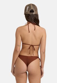 Biquíni castanho com decote halter e calções com laço lateral. Tecido liso, cobertura mínima e ajuste descontraído. Inclui também um boné de rede castanho.