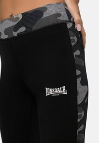 Schwarze Leggings mit einem grauen Camouflage-Muster am Bund, versehen mit einem weißen Logo "Lonsdale London" und strukturierten Seitenpaneelakzenten.