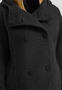 Zwarte textuur peacoat met een dubbelgeknoopte voorkant, grote revers en twee zwarte knopen. Heeft een capuchon en zijzakken.
