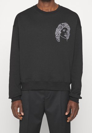 Sweat-shirt noir à manches longues avec un dessin en ligne blanche d'un buste classique sur le haut gauche de la poitrine porté par une personne debout.