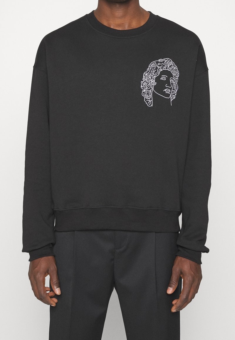 Sweat-shirt noir à manches longues avec un dessin en ligne blanche d'un buste classique sur le haut gauche de la poitrine porté par une personne debout.