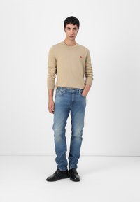 Beige neule, jossa on pyöreä kaula-aukko ja pieni punainen logo. Vaaleansiniset haalarit, joissa on haalistunut ilme. Mustat nahkakengät, joissa on sileä pinta. Minimalistinen muotoilu.
