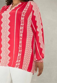 Rote Bluse mit weißen geometrischen und floralen Mustern, langen Ärmeln mit elastischen Bündchen und einem kleinen Schlüssel-Ausschnitt. Weicher Stoff.