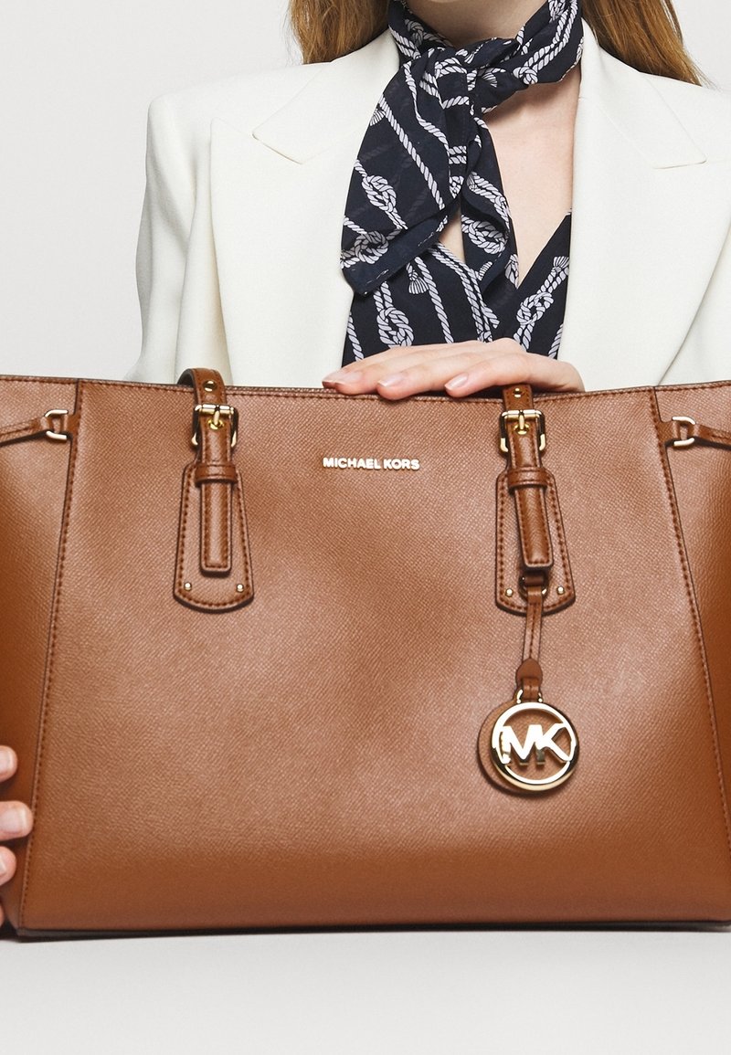 Femme portant un blazer blanc et une écharpe bleu marine tient un grand sac cabas marron Michael Kors avec logo et accents dorés.