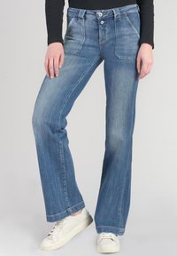 Light blue flared jeans made of denim, featuring a faded wash, dual button closure, and angled front pockets. Worn with white sneakers.