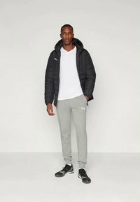 Geacă neagră cu umplutură și glugă, tricou alb, pantaloni de trening gri și pantofi sport negri, având un logo pe geacă și pantaloni.