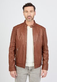 Veste en cuir marron avec un col montant, détails matelassés aux épaules et zips à la taille, portée avec une chemise de couleur claire.