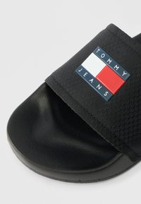 Black slide sandal with textured strap featuring a Tommy Jeans logo patch in red, white, and navy blue colors.