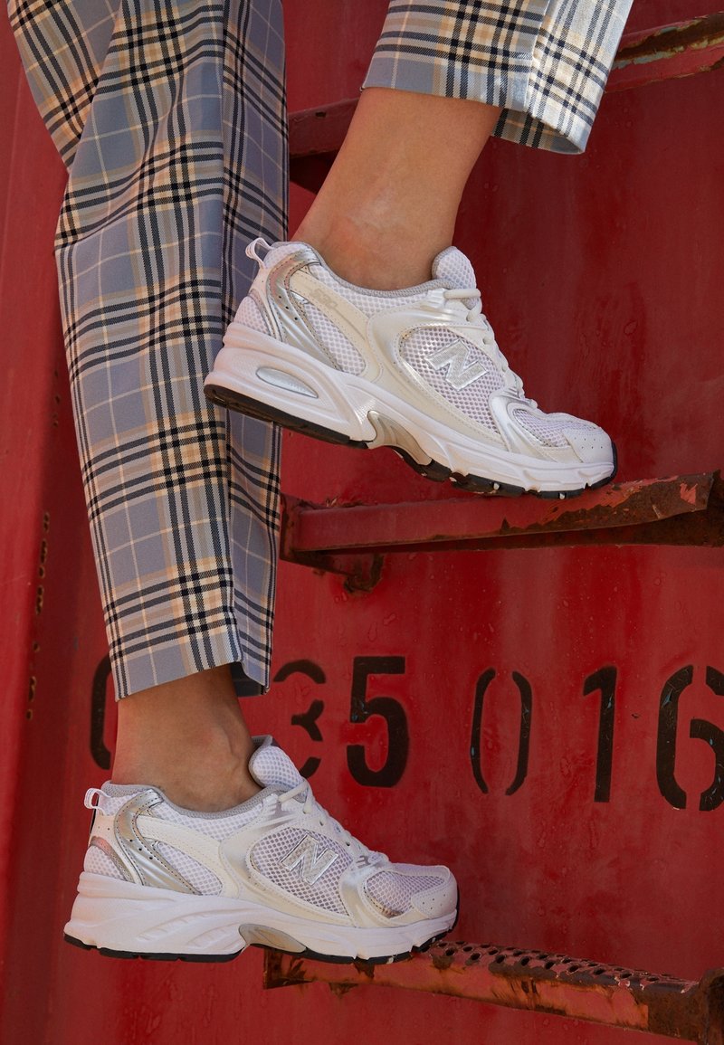 Vita nät-sneakers med reflekterande detaljer och en dämpad sula, kombinerade med rutiga byxor, stående på en röd plattform.