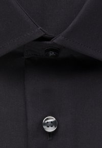 Colletto di camicia nera con il primo bottone allacciato, che mostra la trama del tessuto e i dettagli delle cuciture.