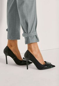 Vrouw in grijze broek met gespen en zwarte hoge hakken met strikaccenten op een lichte achtergrond.