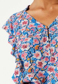 Blouse fleurie avec un décolleté en V, manches courtes à volants, et motifs colorés de fleurs roses et bleues sur un fond clair. 4 boutons visibles.