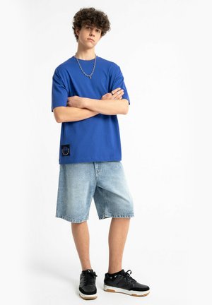 Młody mężczyzna z kręconymi włosami stoi ze skrzyżowanymi ramionami, ubrany w niebieską koszulkę, jeansowe szorty, czarne trampki i łańcuszkowy naszyjnik.
