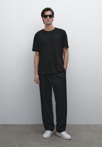 Schwarzes T-Shirt mit kurzen Ärmeln, kombiniert mit lockeren schwarzen Hosen. Tragen mit Sonnenbrille und weißen Sneakern. Glatte Textur, minimalistisches Design.