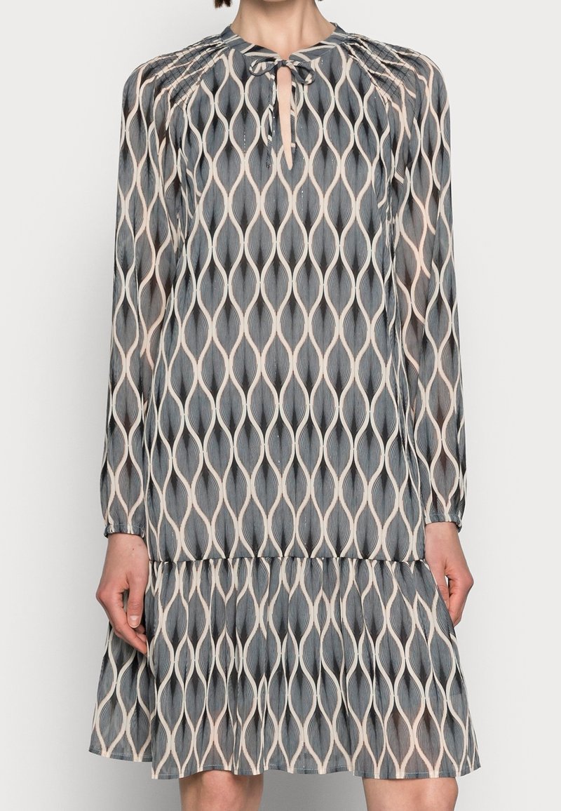 Femme portant une robe mi-longue à manches longues avec un motif ondulé gris, noir et beige, dotée d'une petite encolure goutte d'eau avec un nœud.