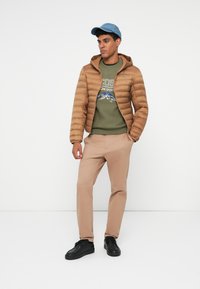 Giacca puffer marrone con cappuccio, maglione verde oliva con stampa grafica, pantaloni beige e scarpe nere. Un cappello di jeans completa l'outfit.