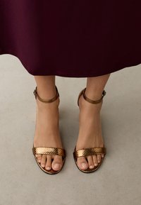 Aranyfémes szandálok, két texturált pánttal és bokapánttal, világos, semleges háttér előtt.