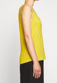 Henkilö, jolla on päällään keltainen ilmanhiha paita ja mustat housut, näkyy profiilissa olkapäästä vyötärölle yksinkertaisen vaalean taustan edessä.