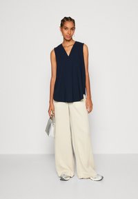 Tunique sans manches bleu marine avec décolleté en V, associée à un pantalon large crème et des accessoires argentés, y compris un petit sac à main.