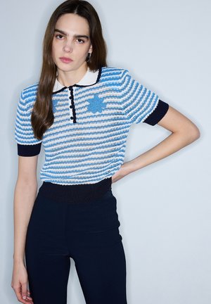 Mujer con cabello largo y castaño que lleva una camisa polo de manga corta de punto a rayas azules y blancas y pantalones oscuros, posando con una mano en la cadera.