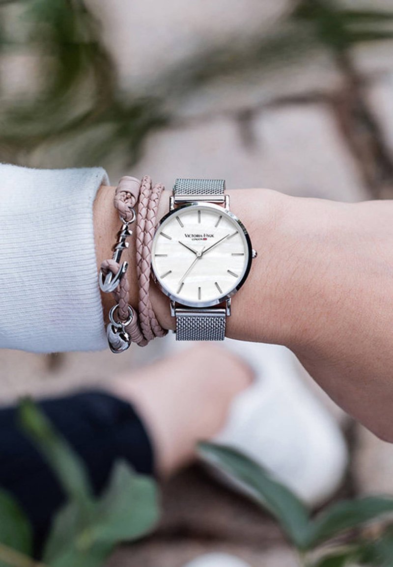 Muñeca con reloj de malla plateada que muestra las 11:26 y pulsera rosa trenzada con charm de ancla, fondo desenfocado con zapatos y vegetación.