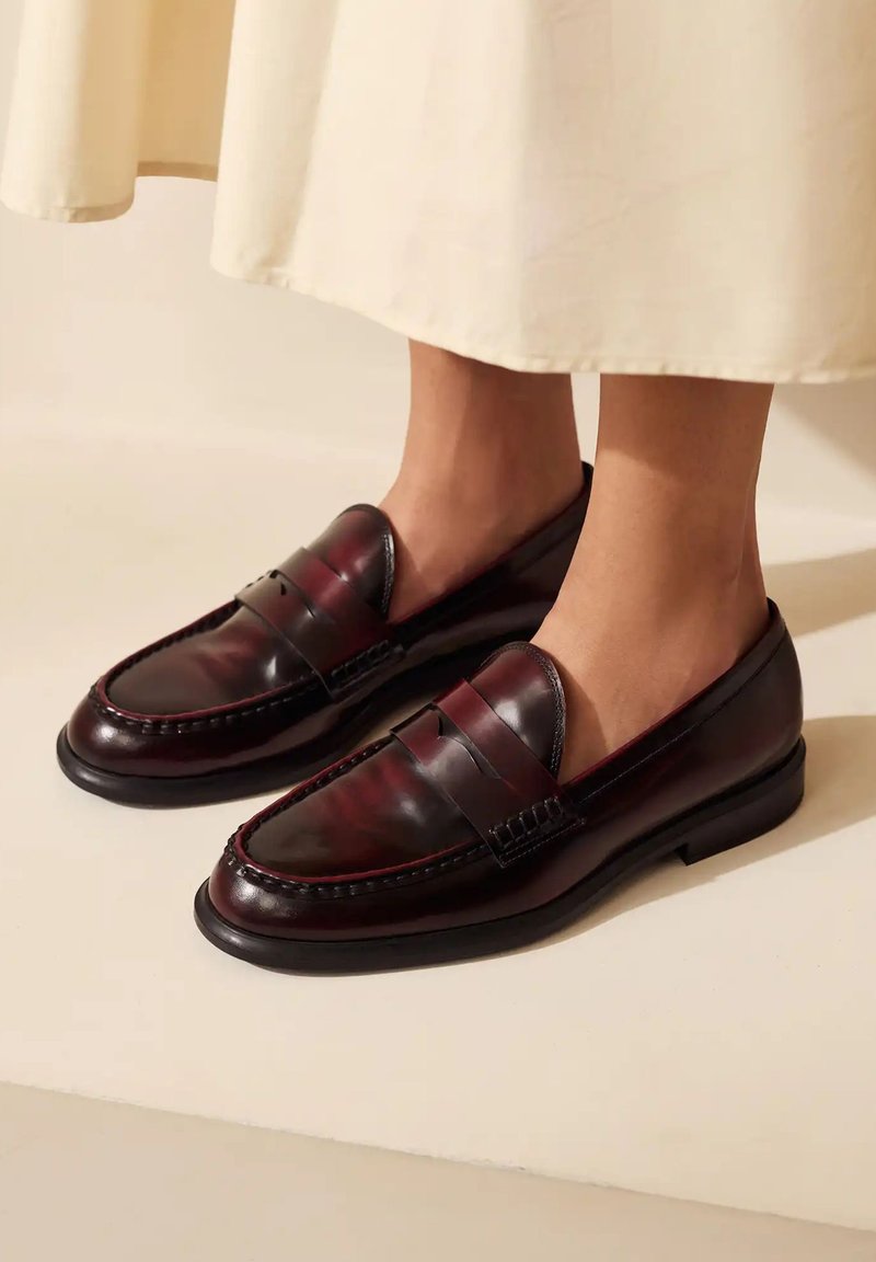 Bordeaux leren loafers met een gladde afwerking, voorzien van een pennyband en gedetailleerde stiksels, rustend op een vlakke zwarte zool.