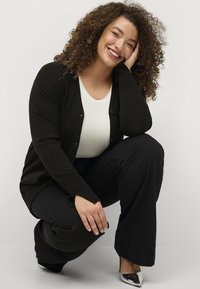 Zwarte cardigan over een witte top, zwarte flare-broek, en zilveren puntige hakken. Krullend haar en een ontspannen houding met een glimlachende uitdrukking.